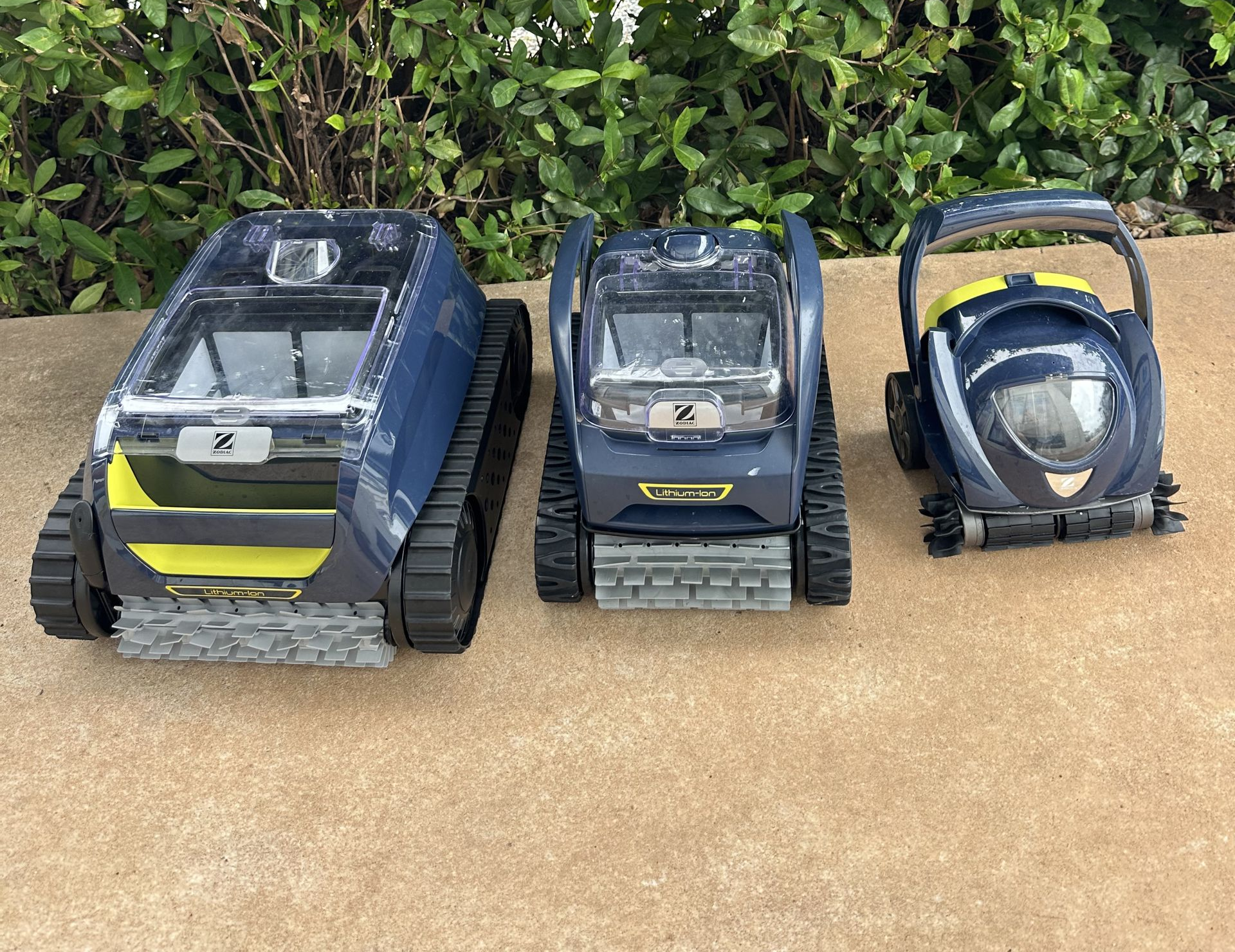 Three robotic pool cleaners, two on the ground and one on a caddy, sit on a concrete path in front of a green hedge — Friendly Pool & Spa Care in Coconut Grove, NT
