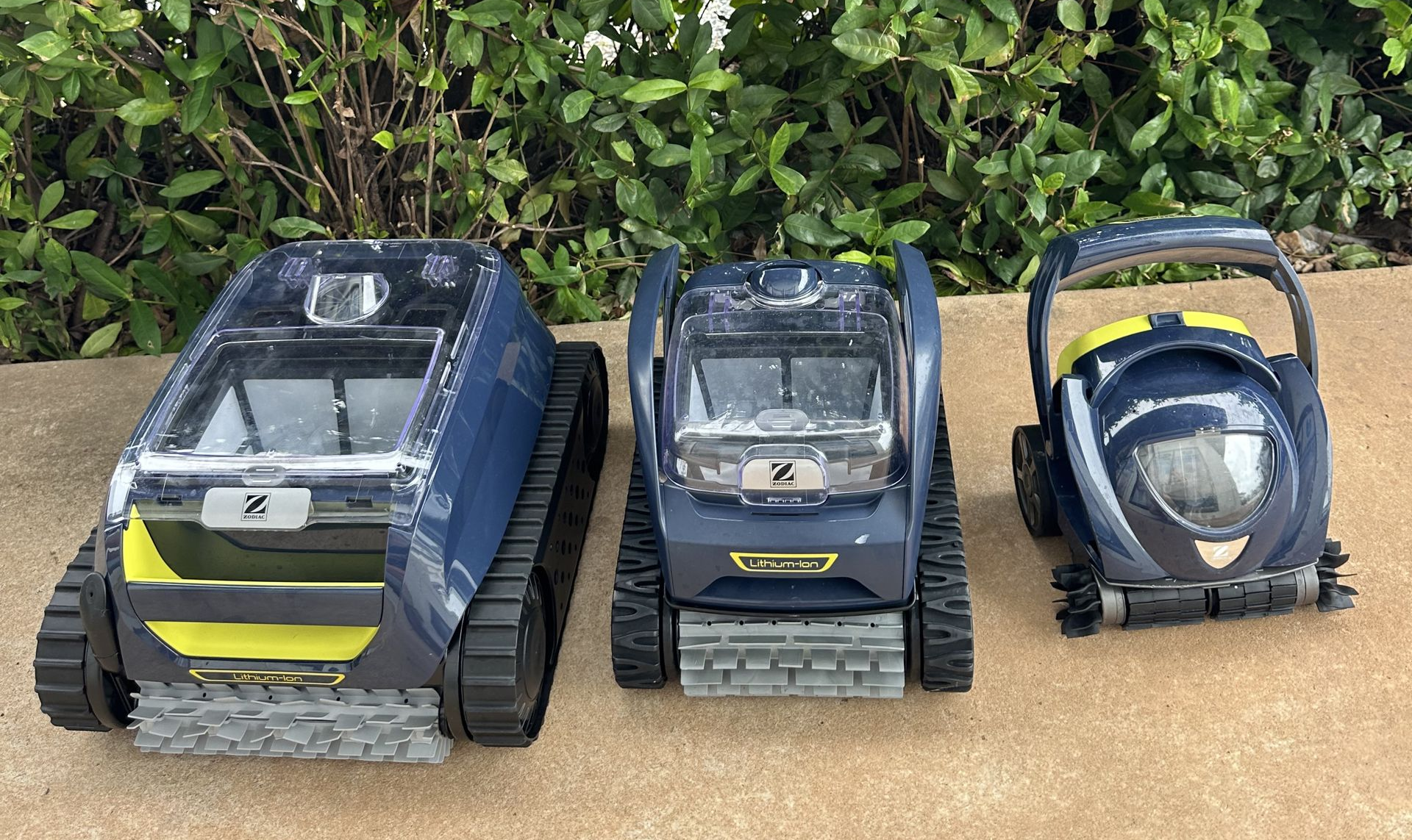 Three robotic pool cleaners of varying sizes lined up on a concrete surface in front of a green hedge  — Friendly Pool & Spa Care in Larrakeyah, NT 