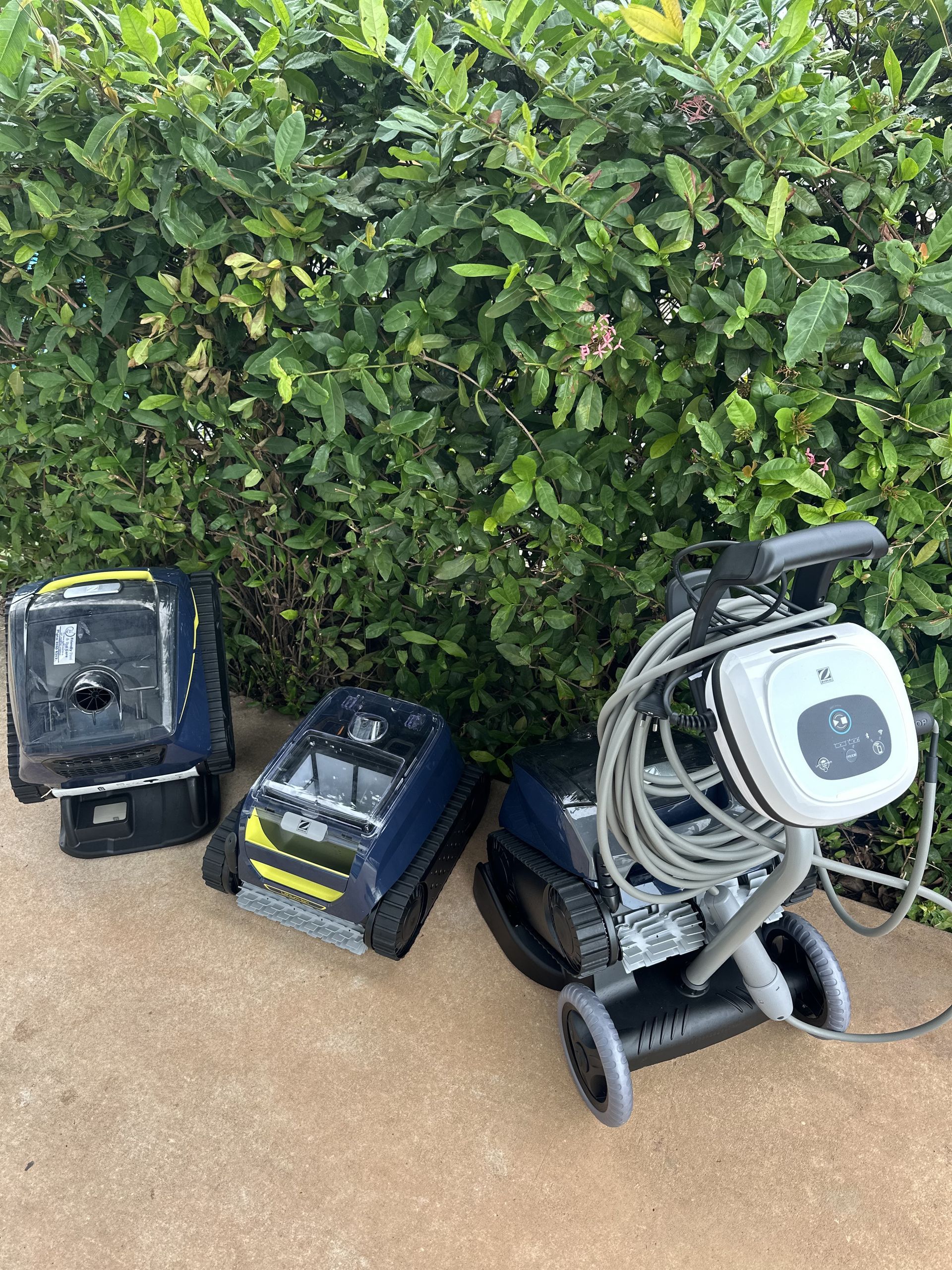 Three automatic pool cleaners in dark blue and yellow, one resting on a transport caddy, arranged on a patio by a hedge — Friendly Pool & Spa Care in Palmerston, NT