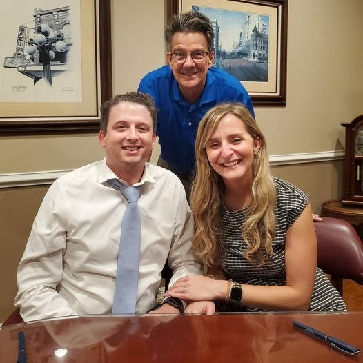 Three people smiling in an office setting. Two are seated at a desk, one standing behind them.
