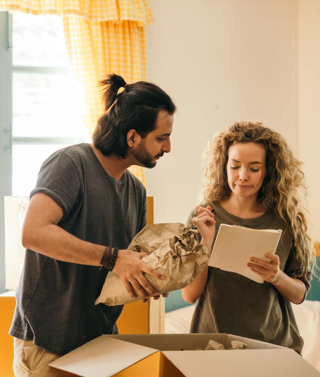 Couple unpacking box, man holding wrapped item, woman reading checklist in bright room.