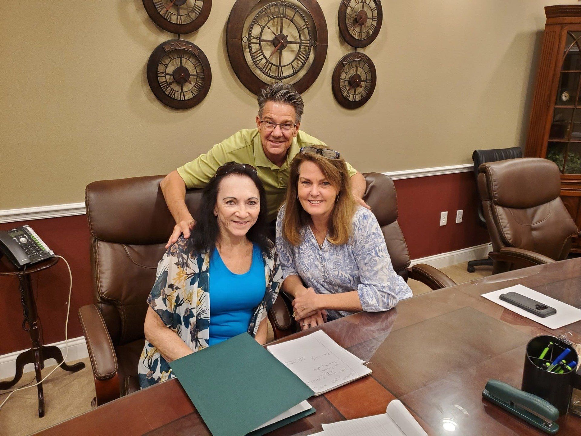 Three people in an office. Two women seated, smiling, with a man standing behind them. They are near a desk with paperwork.