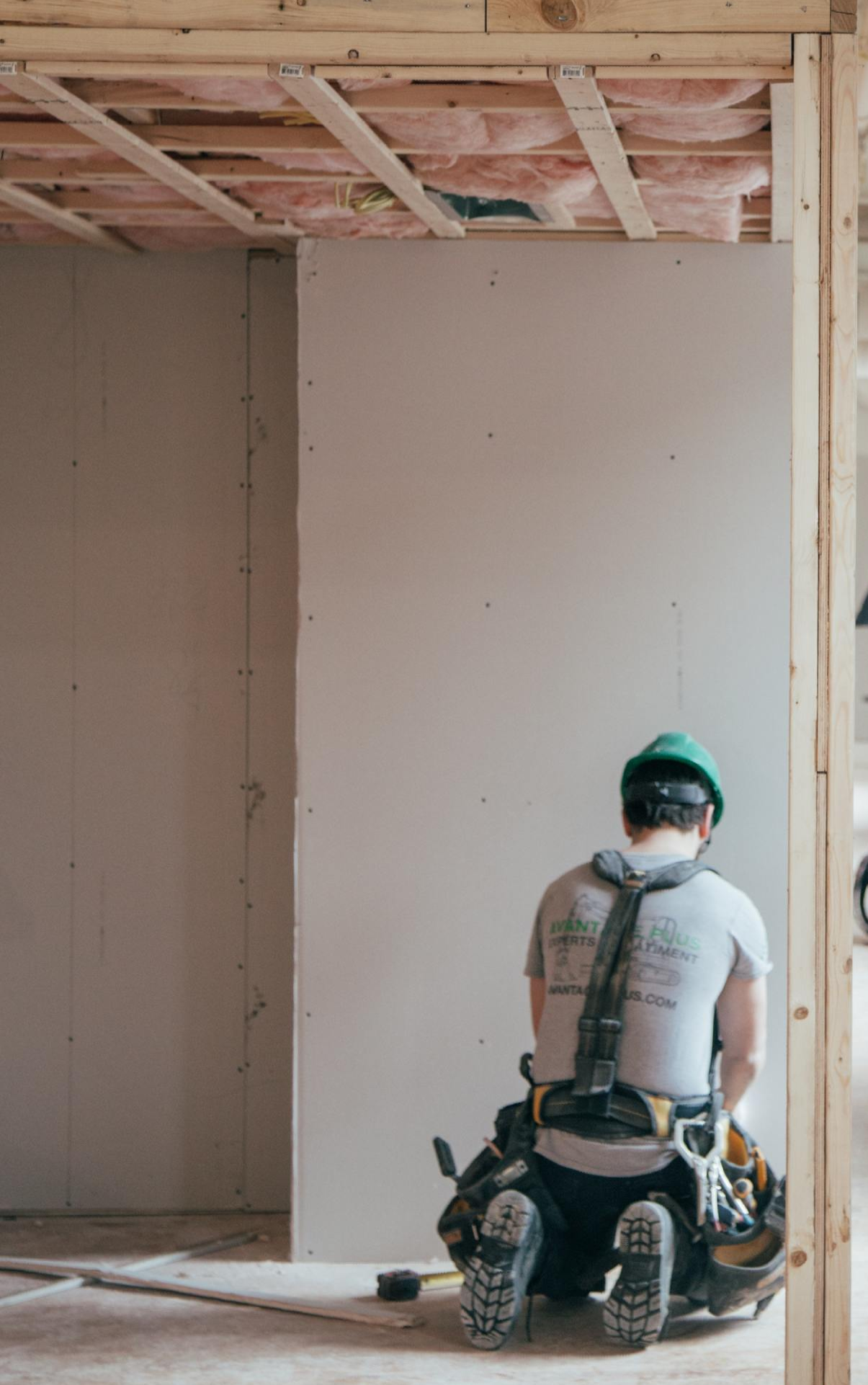 Construction worker installing drywall. Kneeling, wearing a hard hat, and tool belt.