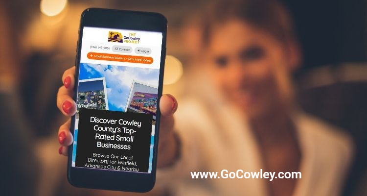 Visit GoCowley.com to find businesses in Ark City and Winfield Kansas A hand holds a smartphone displaying a business directory website for Cowley County against a blurred background.