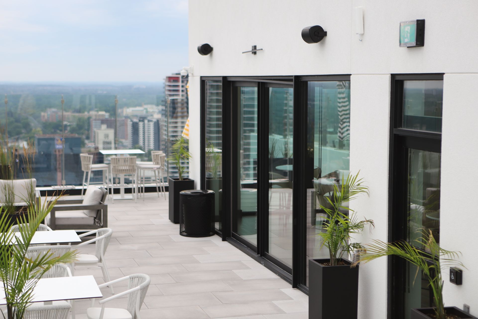 Rooftop patio with city view: white tables, chairs, glass doors, speakers, and potted plants.