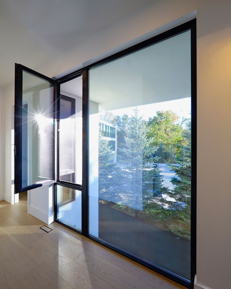 Large, modern window, one panel open. Black frames. Bright sun reflects, trees visible outside. Light interior.