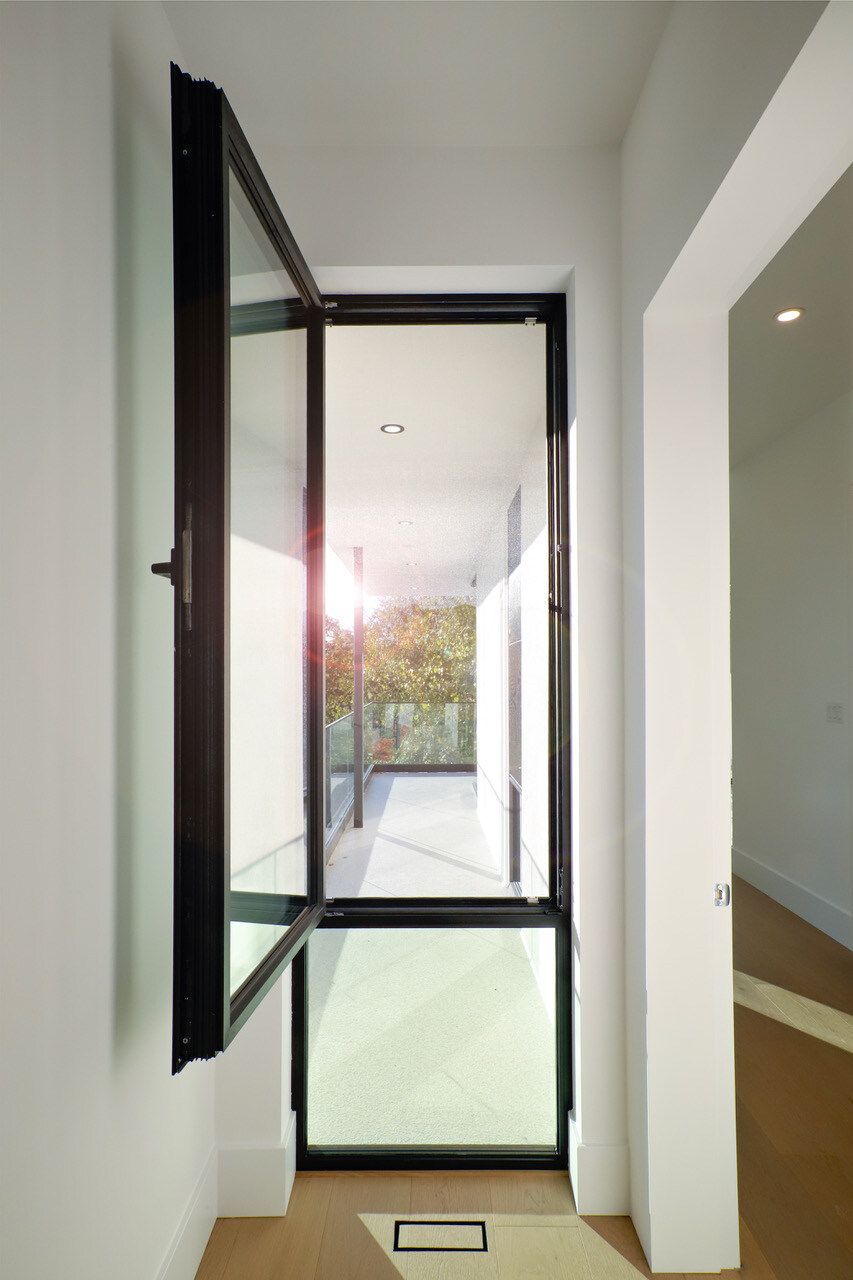 Black-framed glass door ajar, opening to a balcony with greenery. Bright sunlight floods hallway.