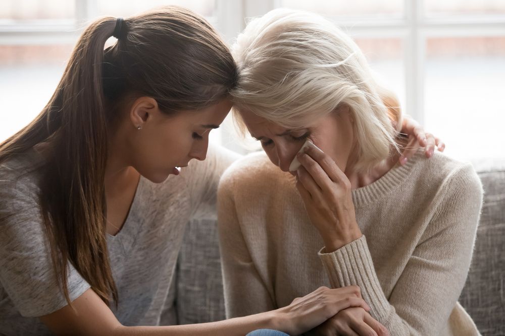 Two women sit closely together, one comforting the other as she wipes away tears.