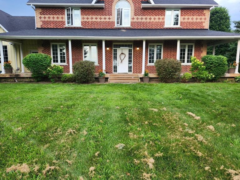 A two-story brick house with a wrap-around porch and dark shingles sits behind a large, green grassy lawn.