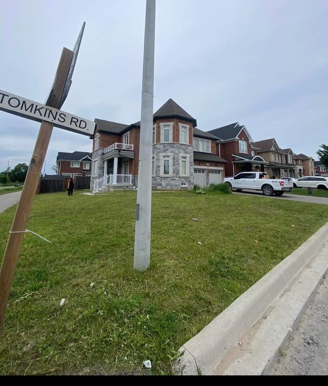 A street-level view shows a suburban two-story brick house with a turret, a lawn, and a street sign labeled Tomkins Rd.