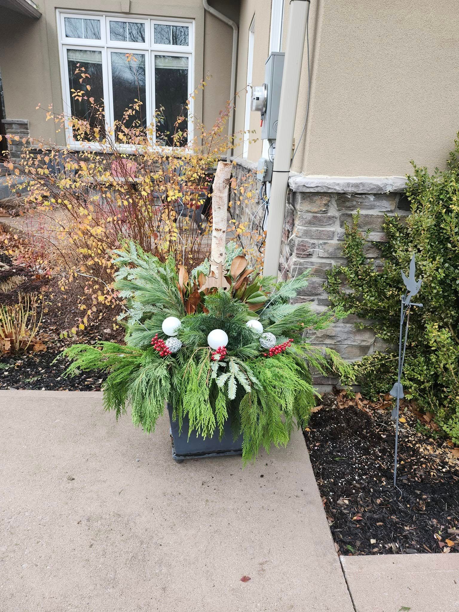 A festive winter planter with evergreen boughs, white ornaments, and red berries sits on a walkway by a home entrance.