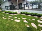 A white house with a deck featuring a black railing, overlooking a lawn with stone pavers and a row of green bushes.