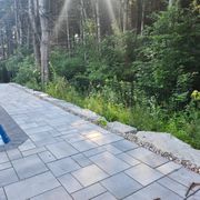 Gray stone pavers form a patio edge next to a pool, separated from a lush, green forest by a line of large rock boulders.