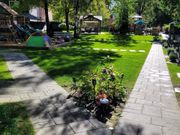 A sunny backyard with two stone pathways, a patch of flowers, a green play tent, and a wooden playground structure.