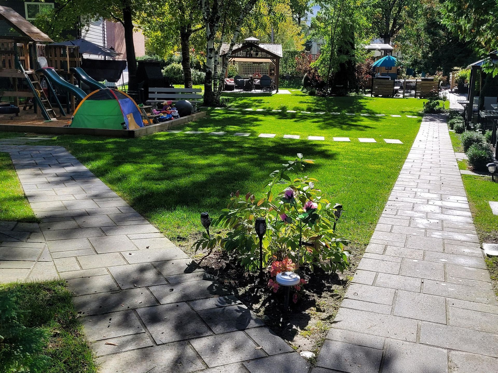 A sunny backyard with two stone pathways, a patch of flowers, a green play tent, and a wooden playground structure.