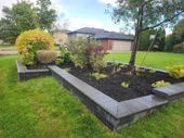 A raised garden bed with dark mulch, various shrubs, and gray stone edging sits in a lush green lawn near a house.
