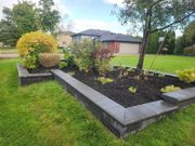 A raised garden bed with dark mulch, various shrubs, and gray stone edging sits in a lush green lawn near a house.