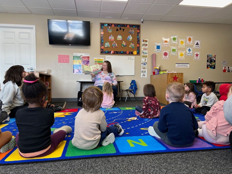 A group of children are sitting on the floor listening to a teacher read a book.