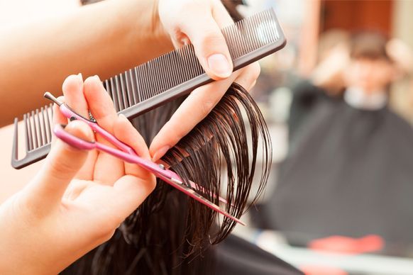 Manos usando tijeras y un peine rosa para cortar cabello oscuro en un salón de belleza.
