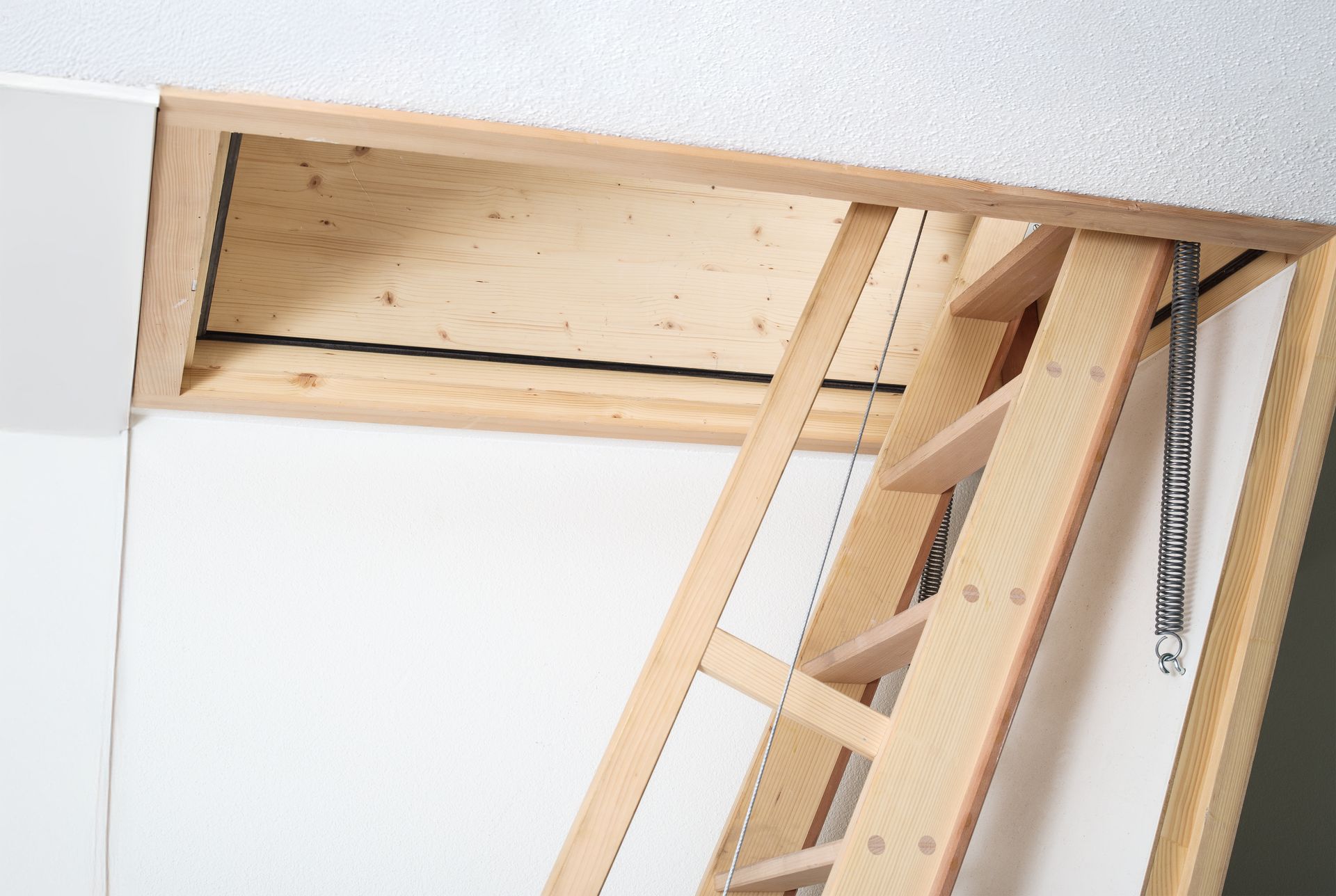 A wooden attic ladder extends downward from a ceiling hatch into a room with white walls.