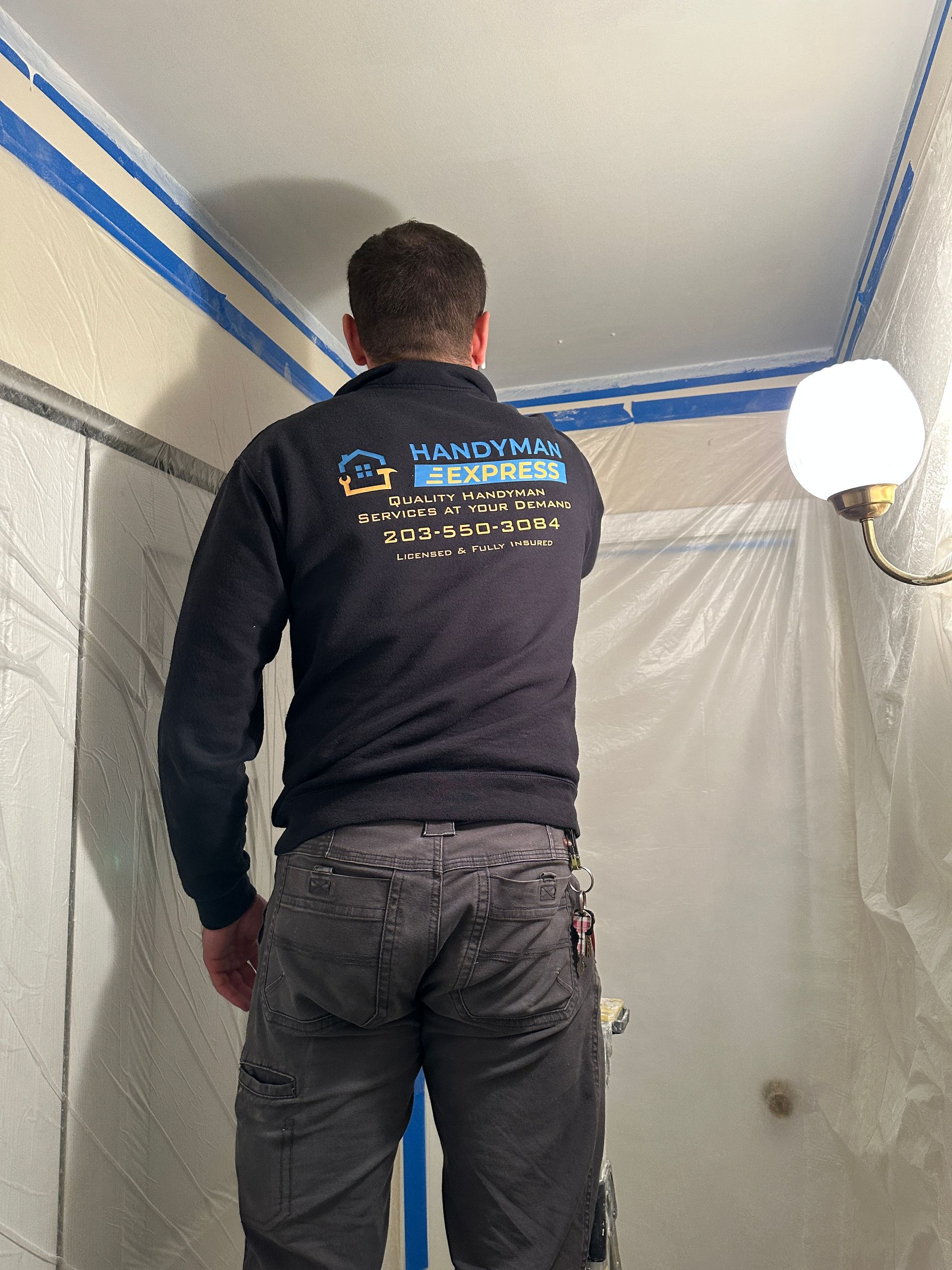 A worker in a branded dark hoodie and grey work pants prepares a ceiling for painting in a room covered with plastic.