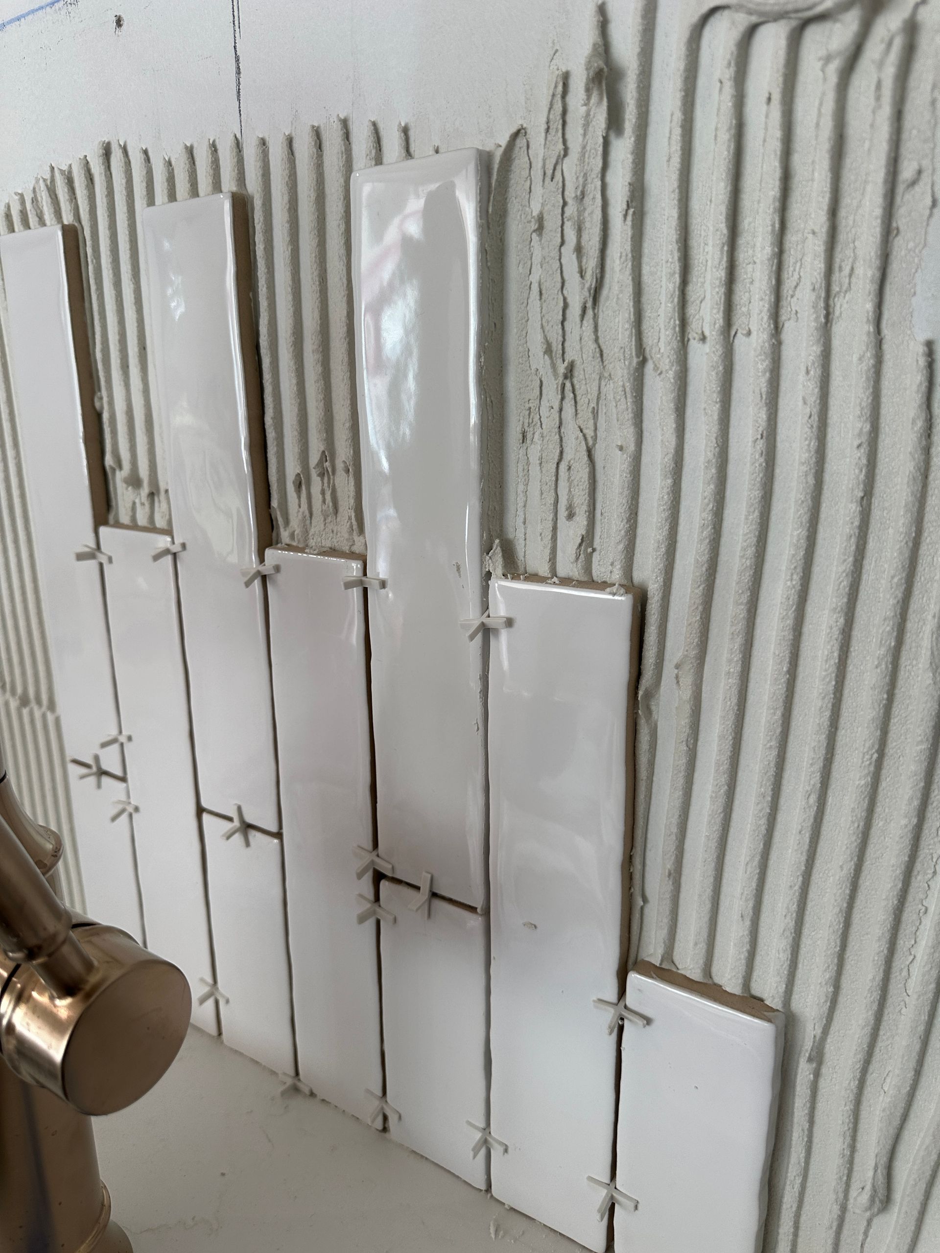 Vertical white subway tiles are being installed on a wall with mortar ridges and plastic spacers visible.