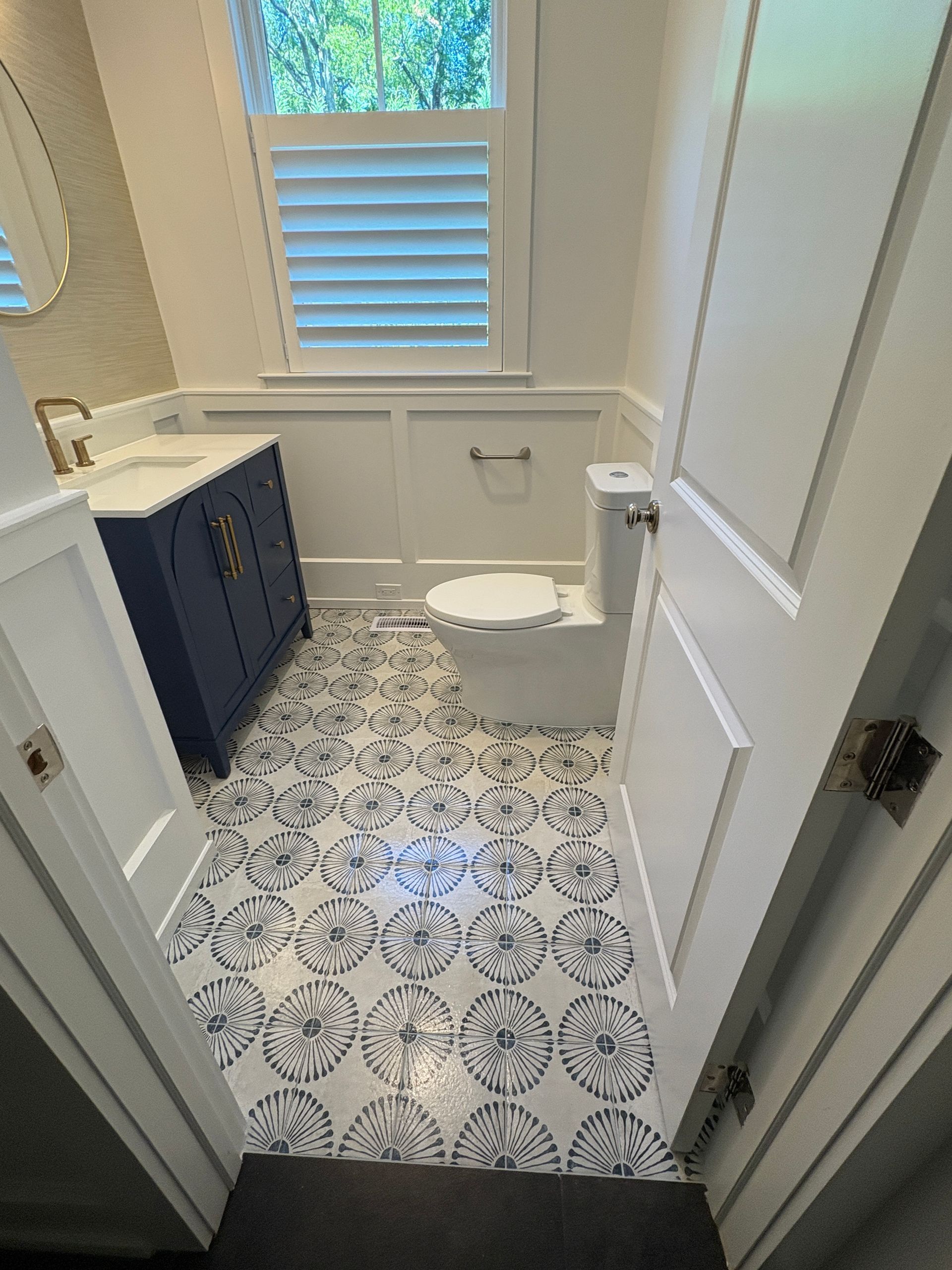 A small bathroom with white wainscoting, a navy blue vanity, a toilet, and patterned blue and white tile floors.