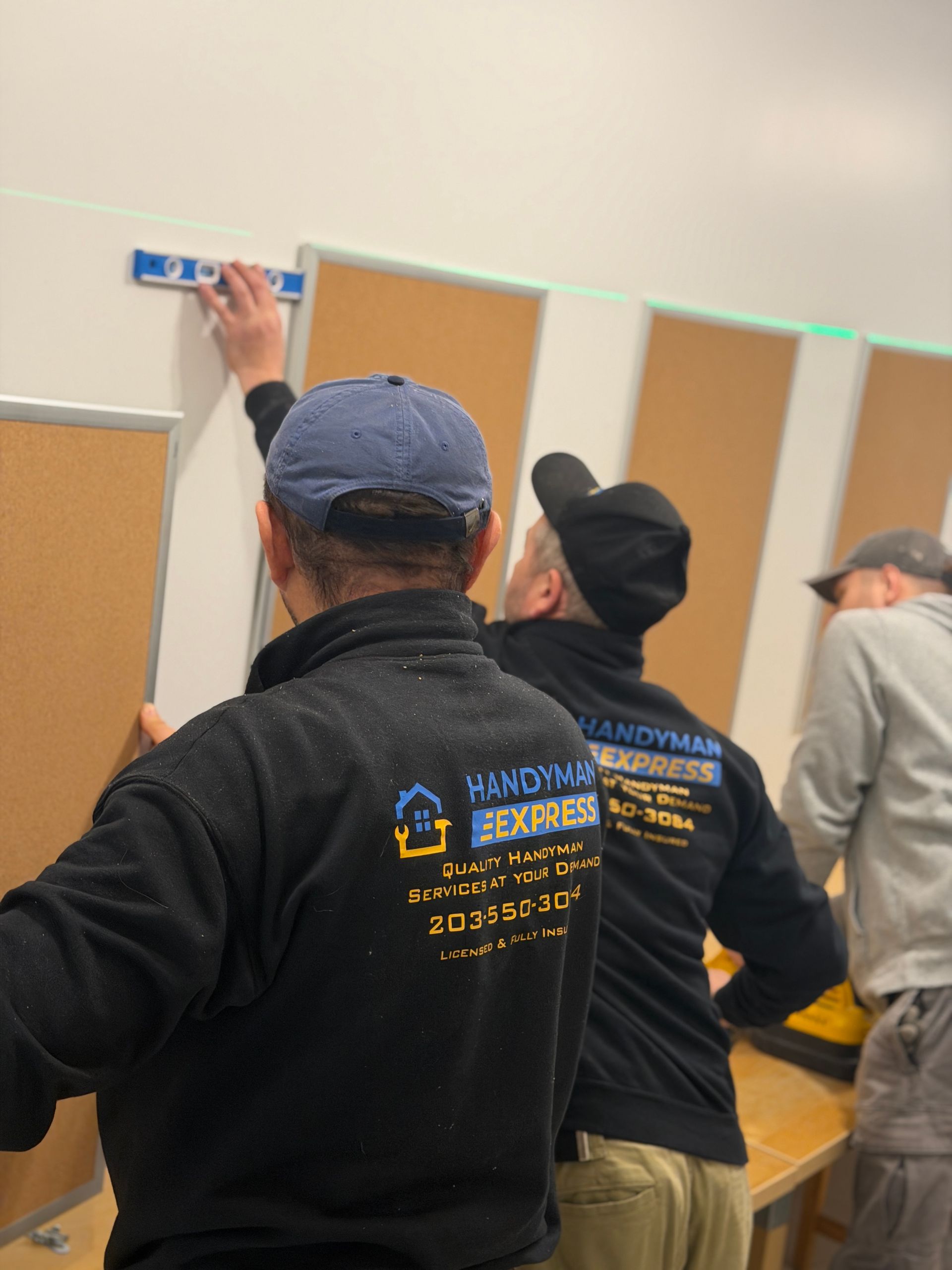 Three workers in uniform install cork boards on a white wall, using a level to ensure they are straight.