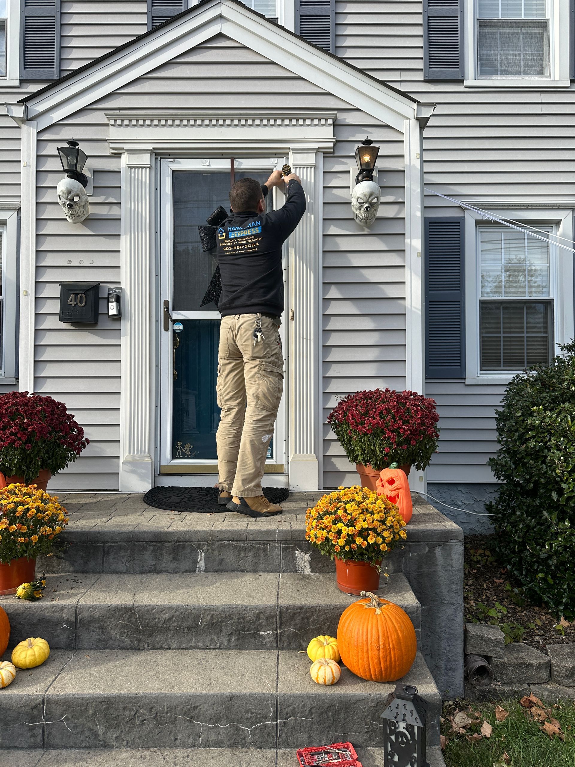 A person standing on a front porch step, installing holiday decorations above a doorway surrounded by autumn decor.