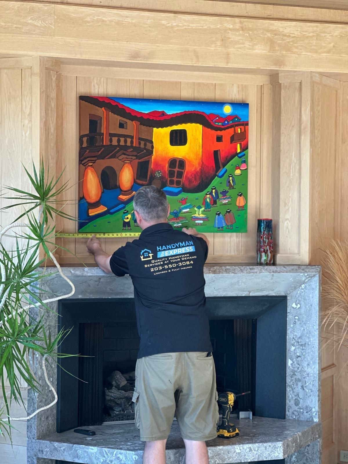 Person hanging a colorful painting above a fireplace in a wood-paneled room