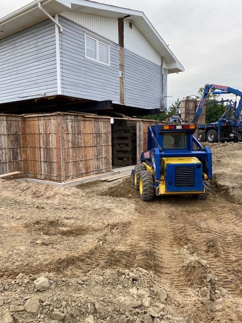 Maison surélevée sur pilotis en bois ; chargeuse compacte bleue et jaune sur terre, grue en arrière-plan.