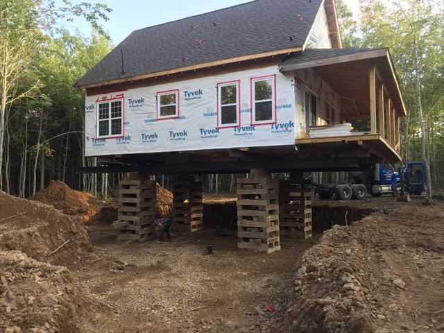 Maison surélevée sur pilotis, en construction dans une zone boisée. Bâche bleue et fenêtres visibles.
