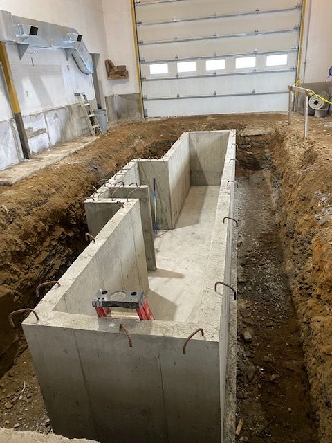 Des fondations en béton sont en cours de construction dans une tranchée creusée à l'intérieur d'un bâtiment, avec une porte de garage fermée en arrière-plan.