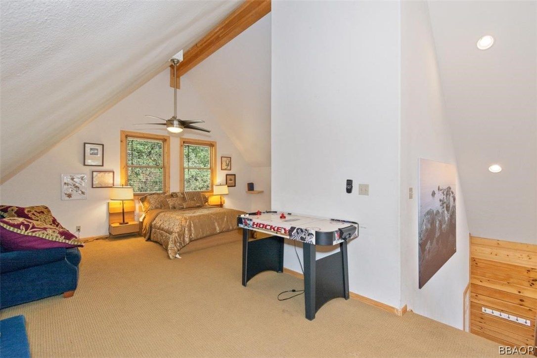 Loft bedroom with bed, armchair, table, and windows. Light walls and ceiling.