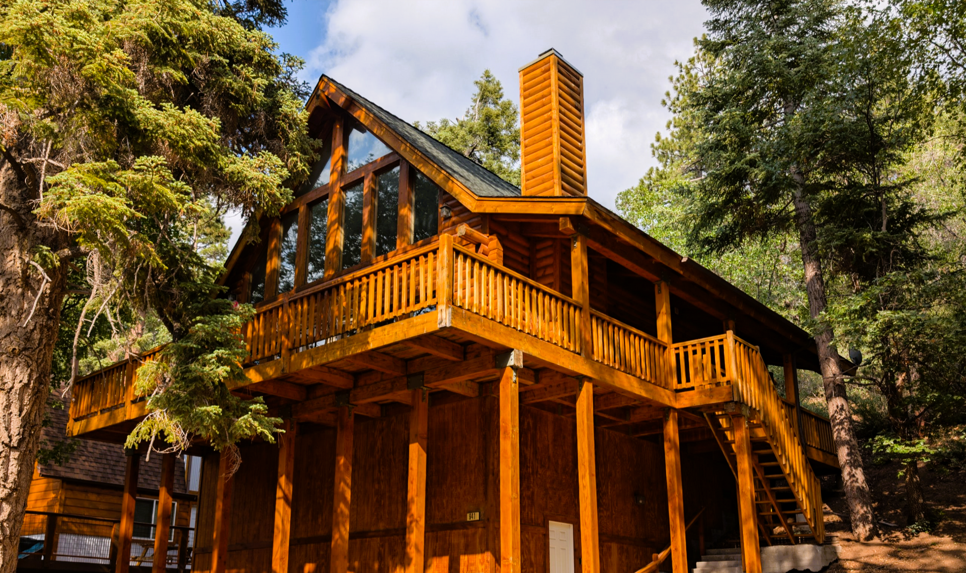 Wooden cabin in a forest, with a porch and chimney, surrounded by trees, sunny day.