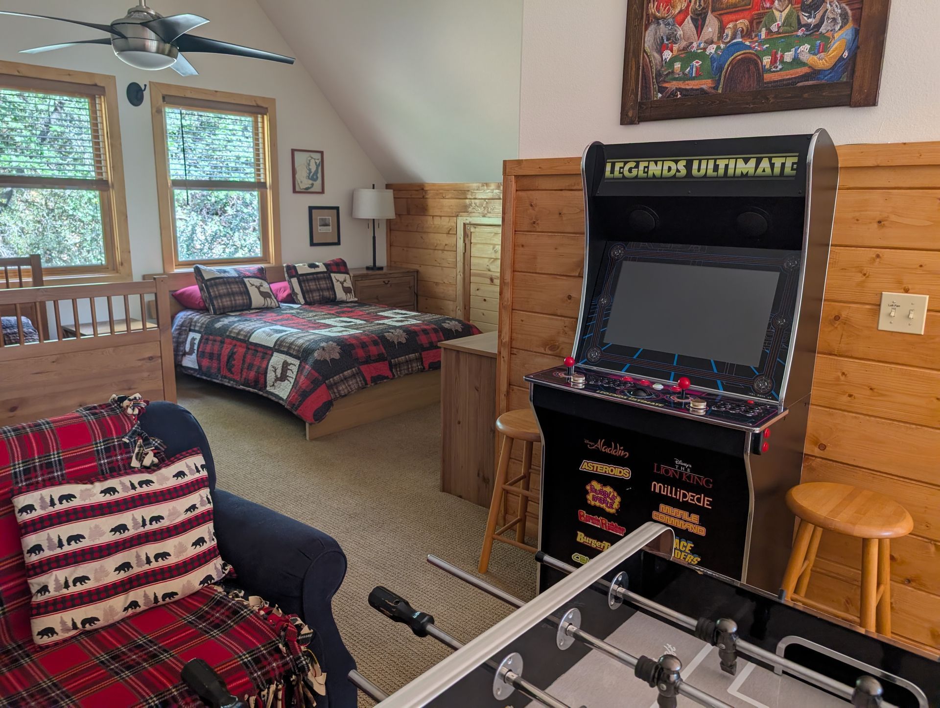 A cozy cabin bedroom with arcade game, foosball table, bed, and sofa. Wooden walls, windows, and bear-themed decor.