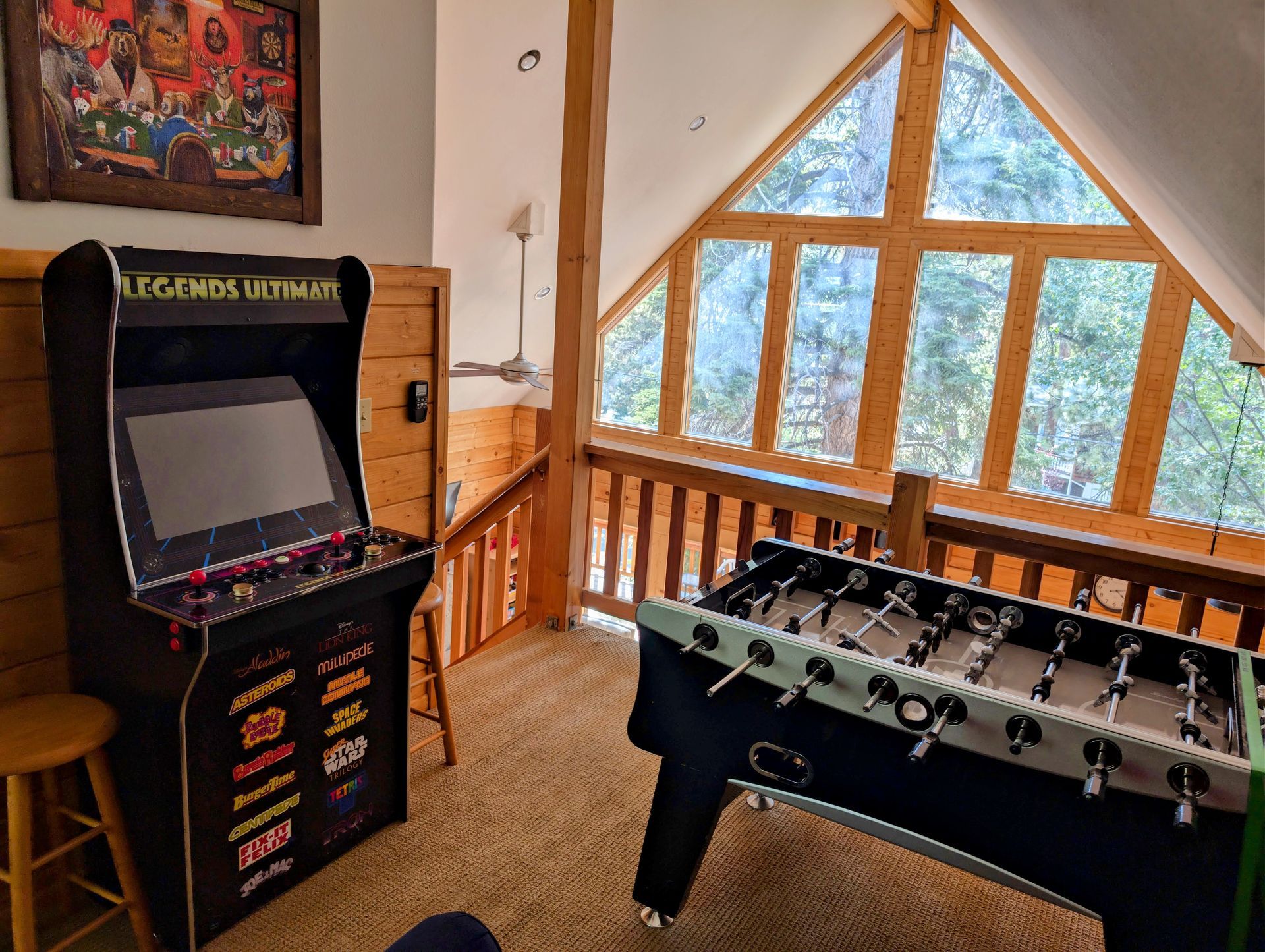 Arcade game and foosball table in a cabin loft with a large window overlooking trees.