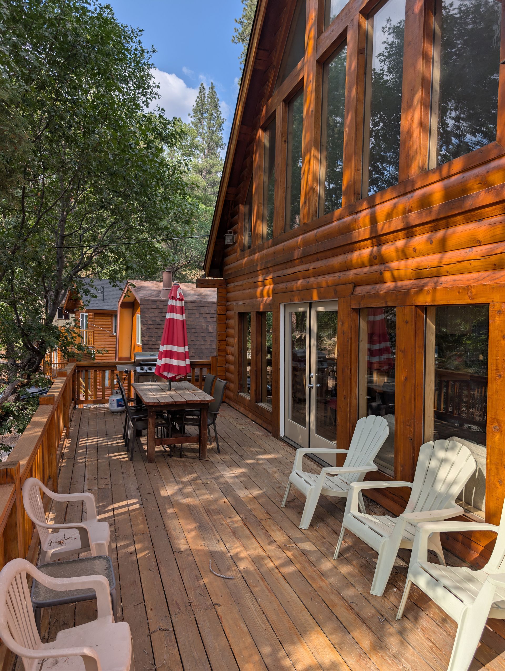 Log cabin with wooden deck, patio furniture, and a red umbrella. Sunlight, trees, and a blue sky.