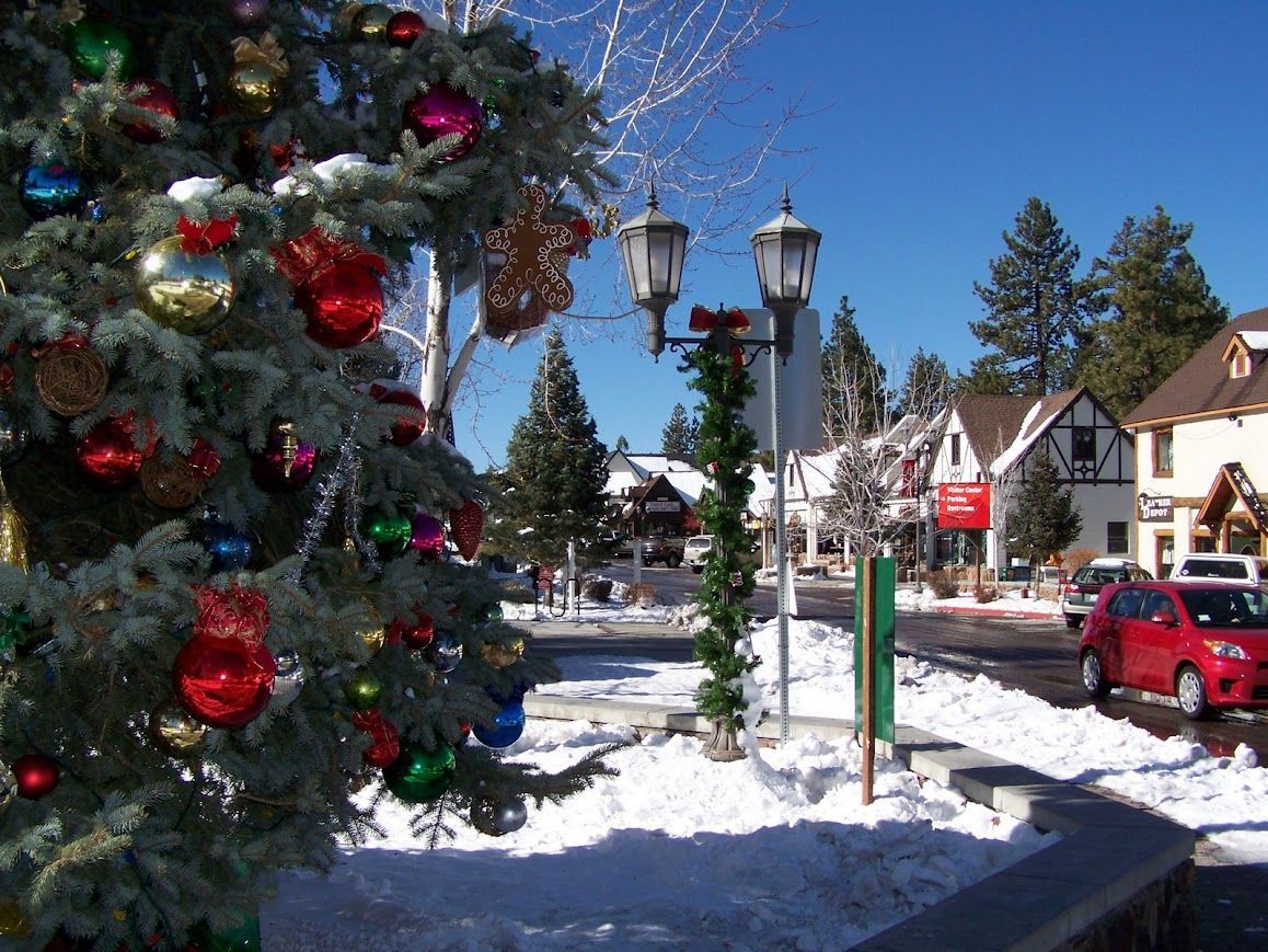 Snowy village square with Christmas tree and decorated lampposts, shops in the background, blue sky.