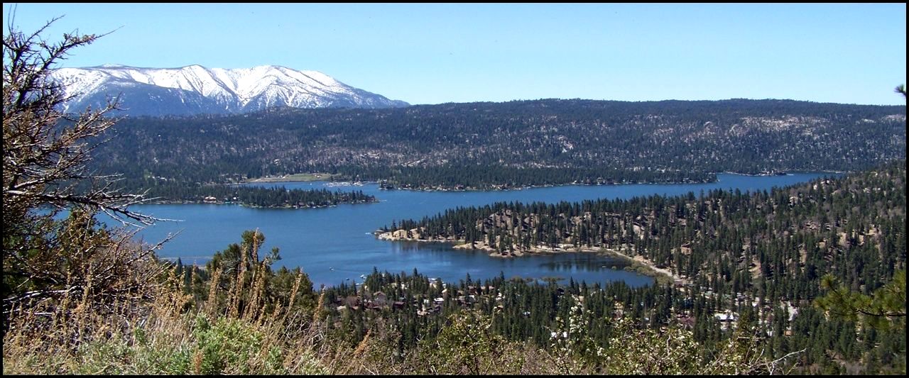 Scenic view of a large lake surrounded by forest, with snow-capped mountains in the background under a blue sky.