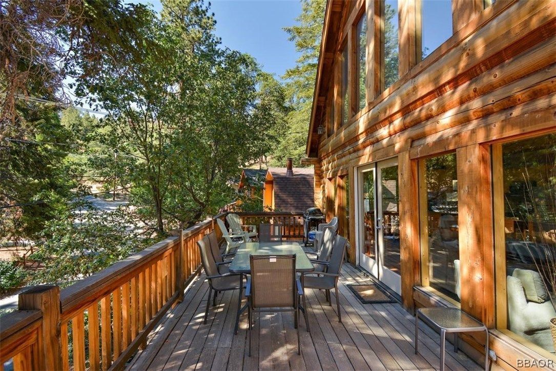 Wooden deck with dining table and chairs outside a cabin with large windows; surrounded by trees.
