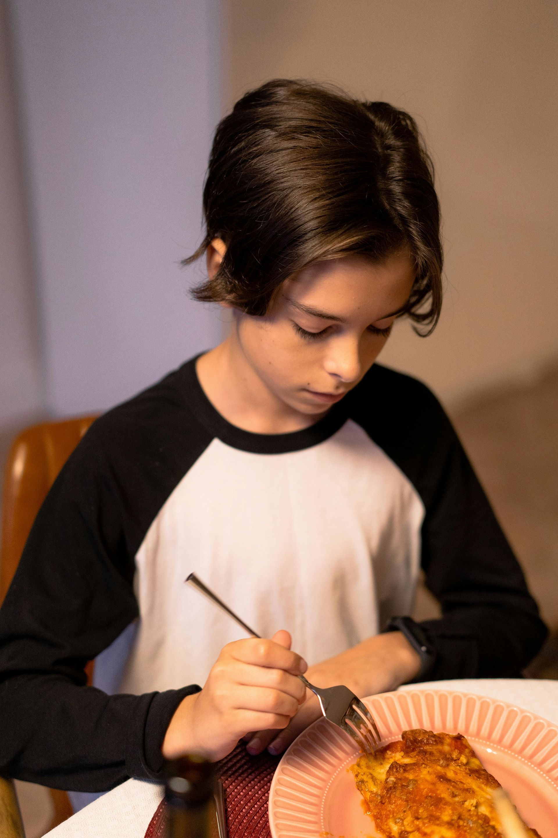 Child eating at a table, looking down while cutting food with a fork and knife.