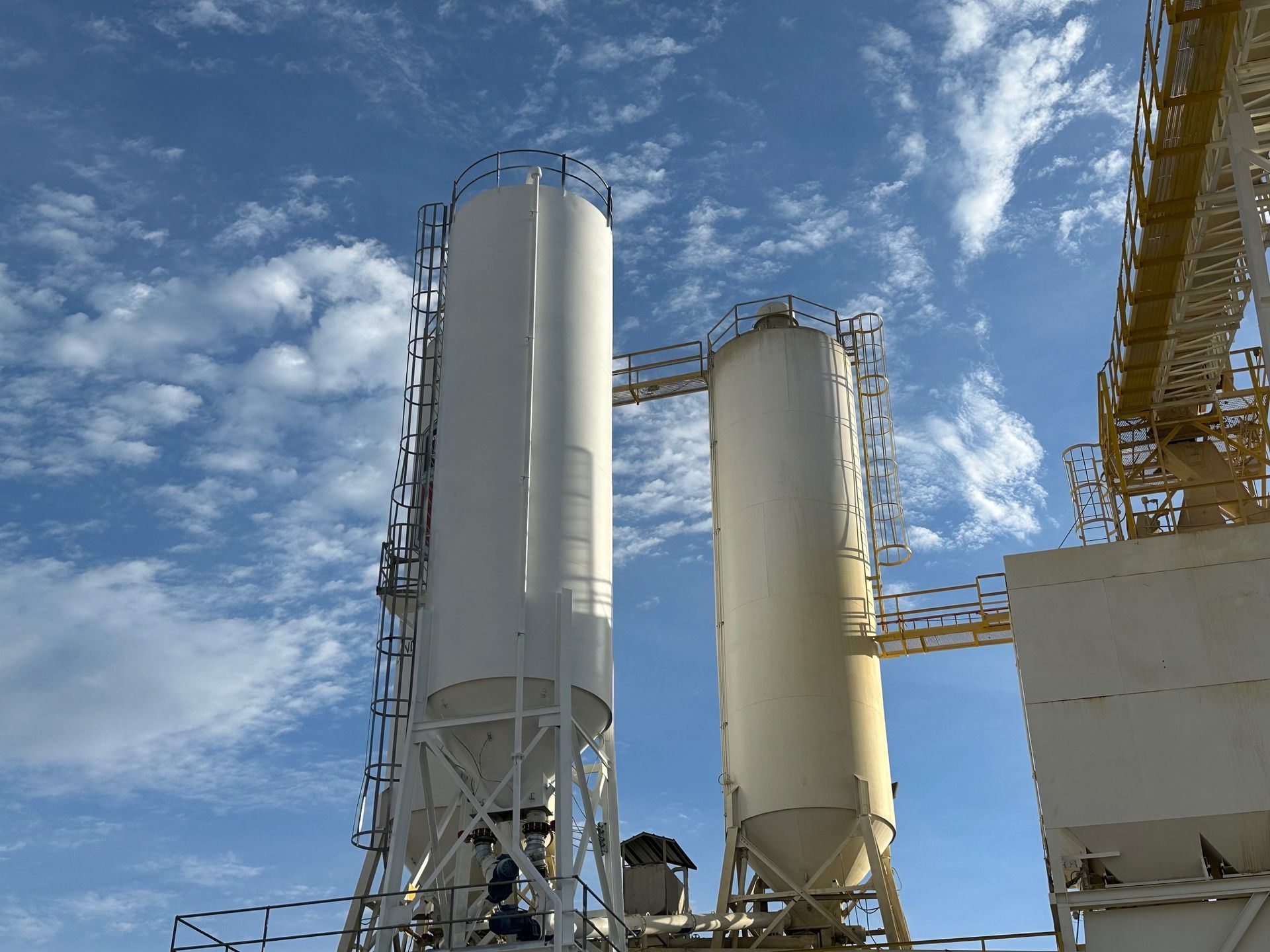 Two tall, white industrial silos against a blue sky with fluffy clouds