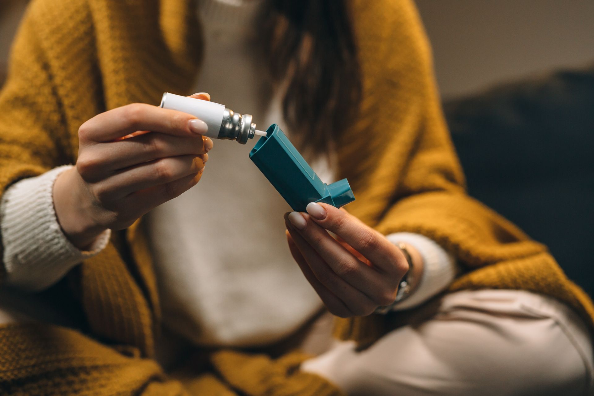 Person opens a teal inhaler beside a white tissue, seated indoors in a mustard sweater