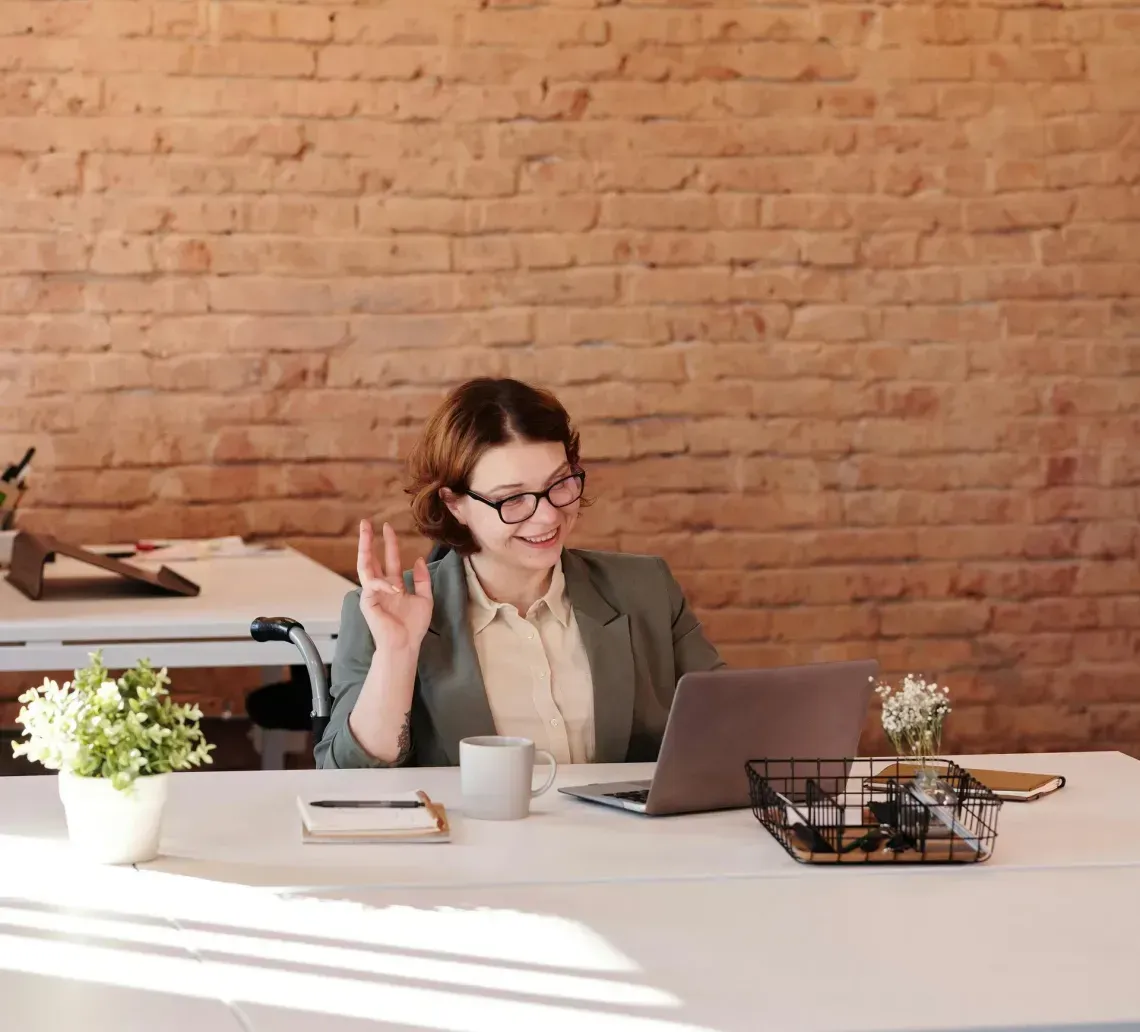 Woman in a wheelchair waves during a video call in an office with brick wall.