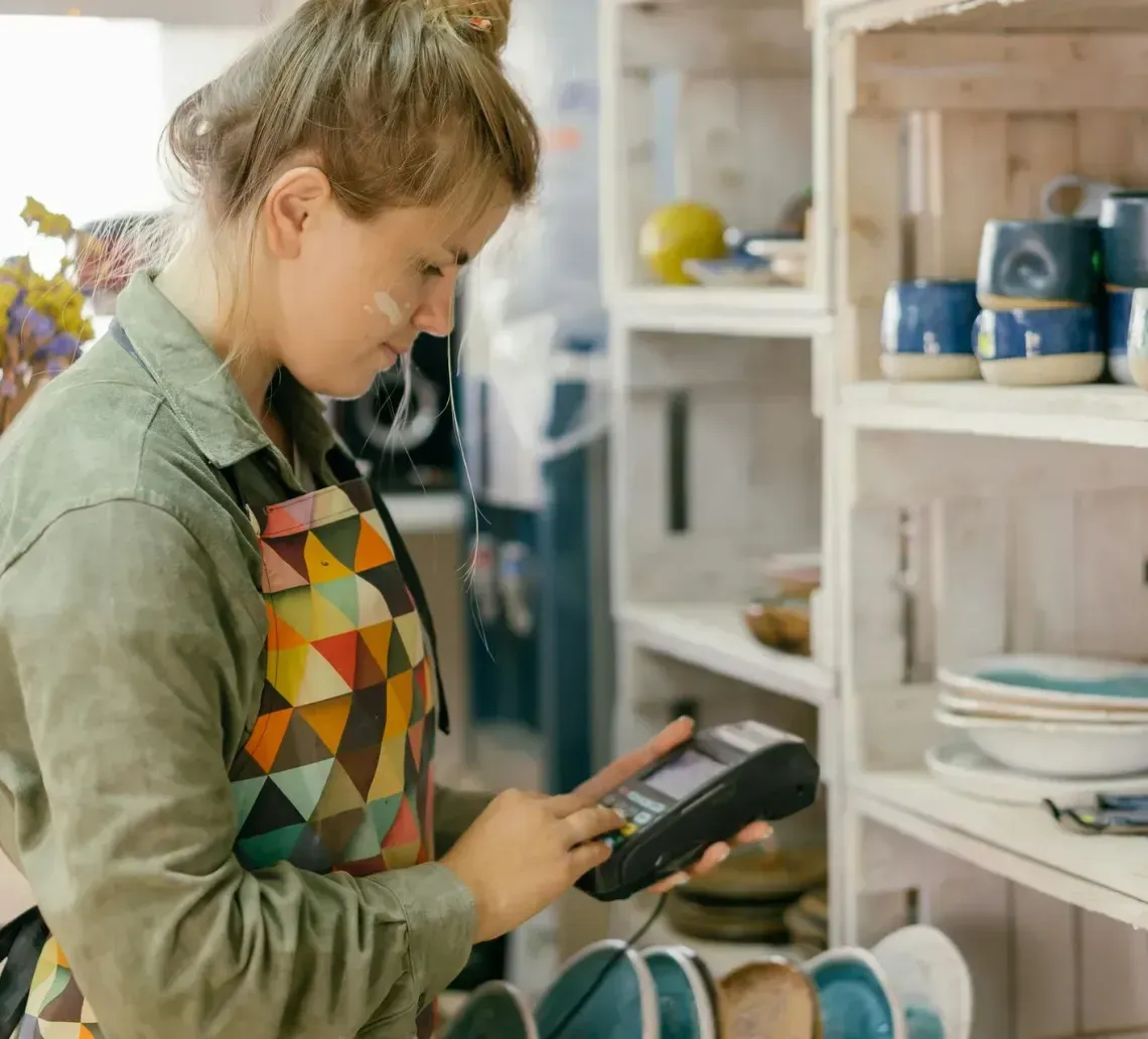 Woman in apron using a payment device in a shop with shelves of ceramic goods.