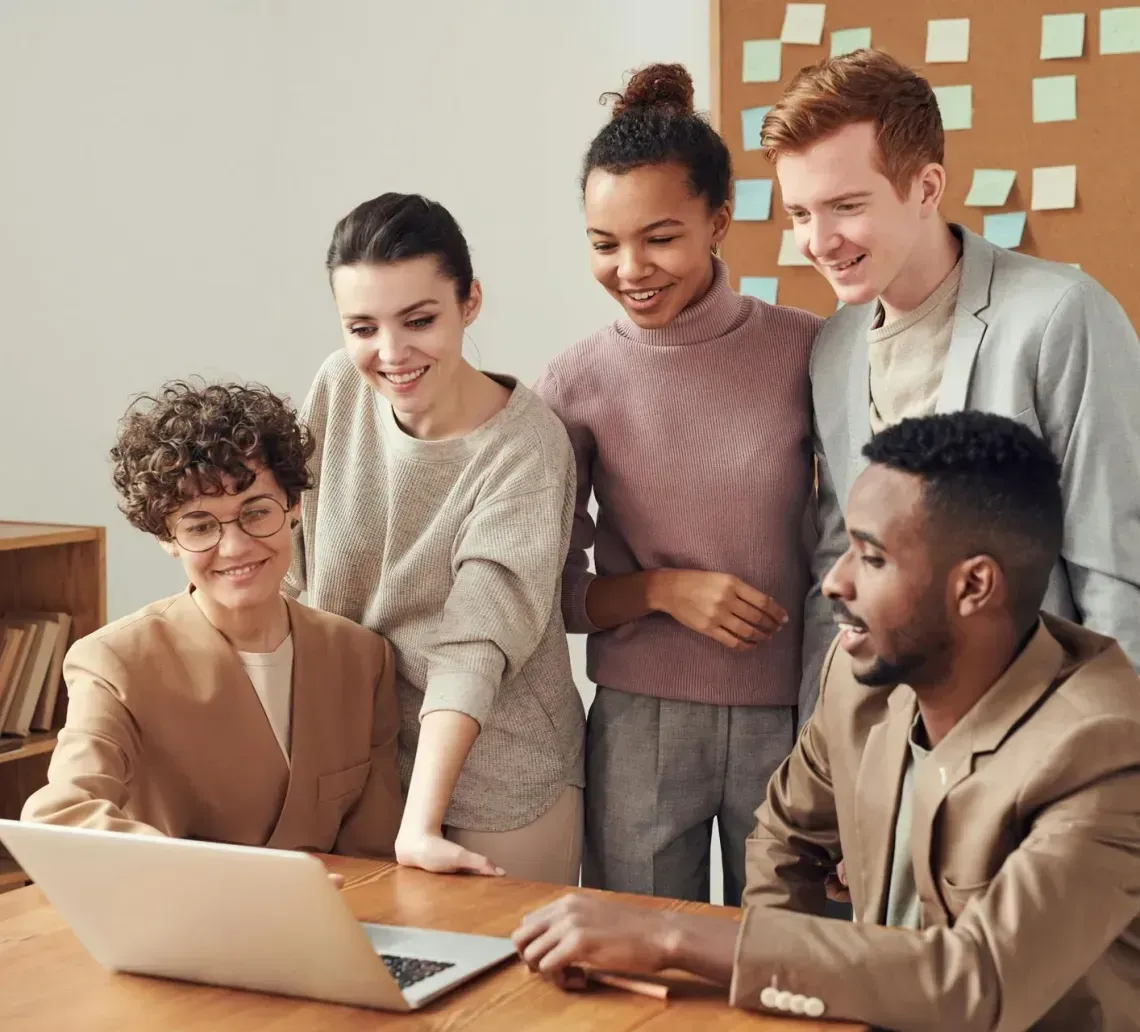 Five coworkers gathered around a laptop, smiling. Light-colored office setting with sticky notes on the wall.