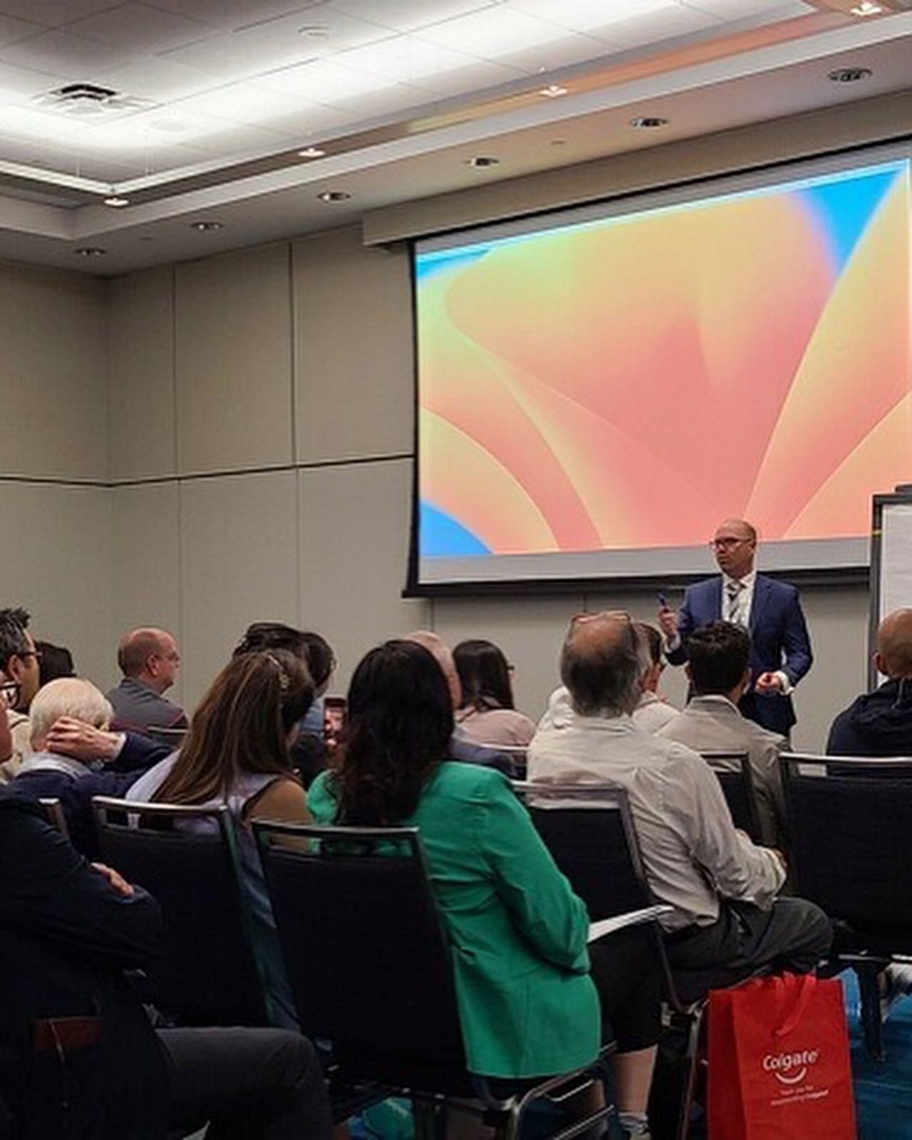 Audience attending a presentation in a conference room, with a speaker beside a large colorful screen.