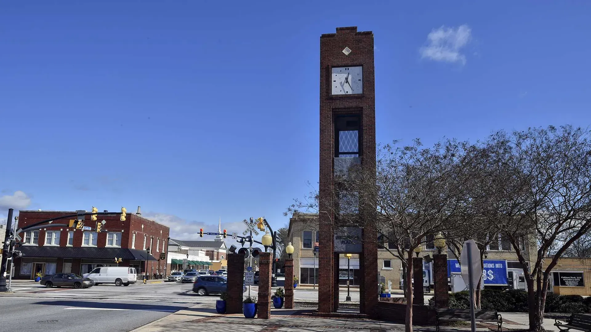Brick clock tower in a snowy downtown square under a blue sky, with buildings and bare trees nearby