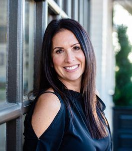 Smiling woman in a black top leaning by a window in a bright hallway